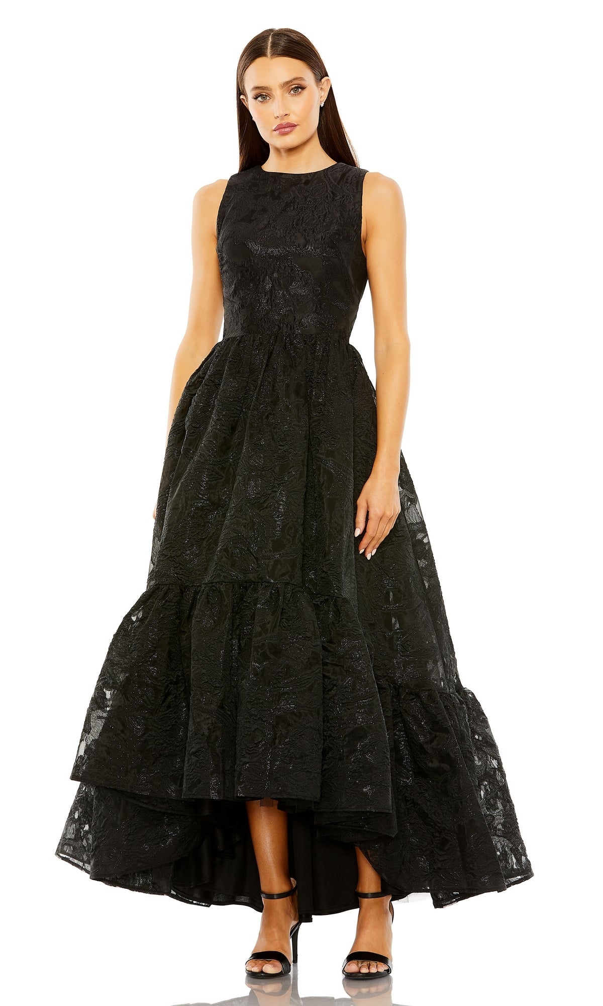 A woman in the Mac Duggal 20714 Brocade High-Low Formal Dress with a black textured fitted bodice and tiered skirt, styled with black high-heeled sandals, stands against a plain background.
